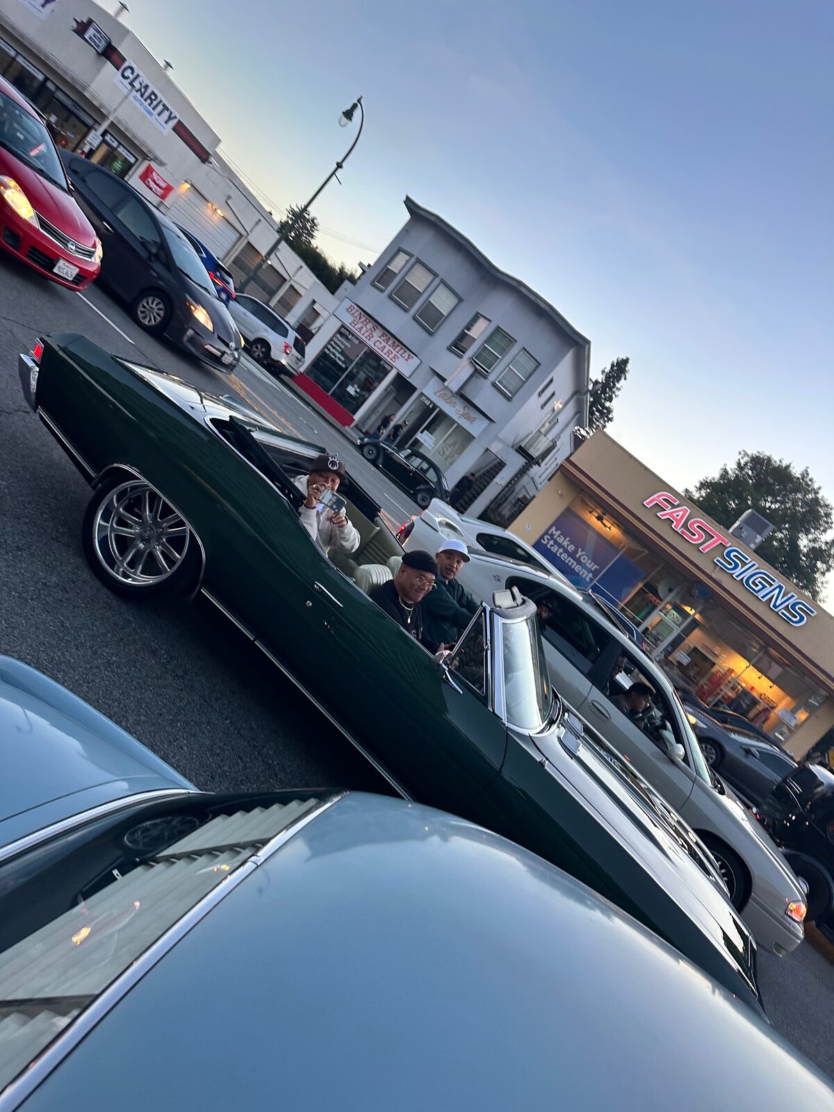 A dark green convertible on El Camino Real at dusk with storefronts behind it