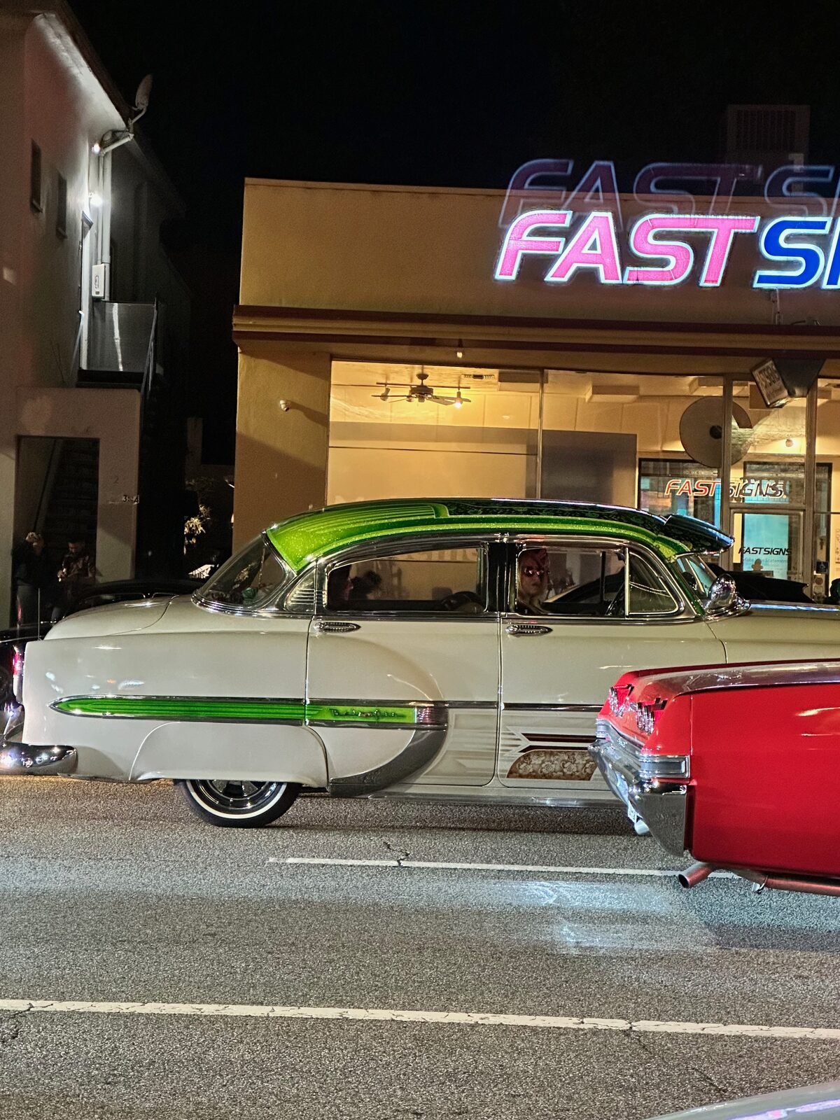 A white 1950s Chevrolet with green scalloped paint parked at night under neon