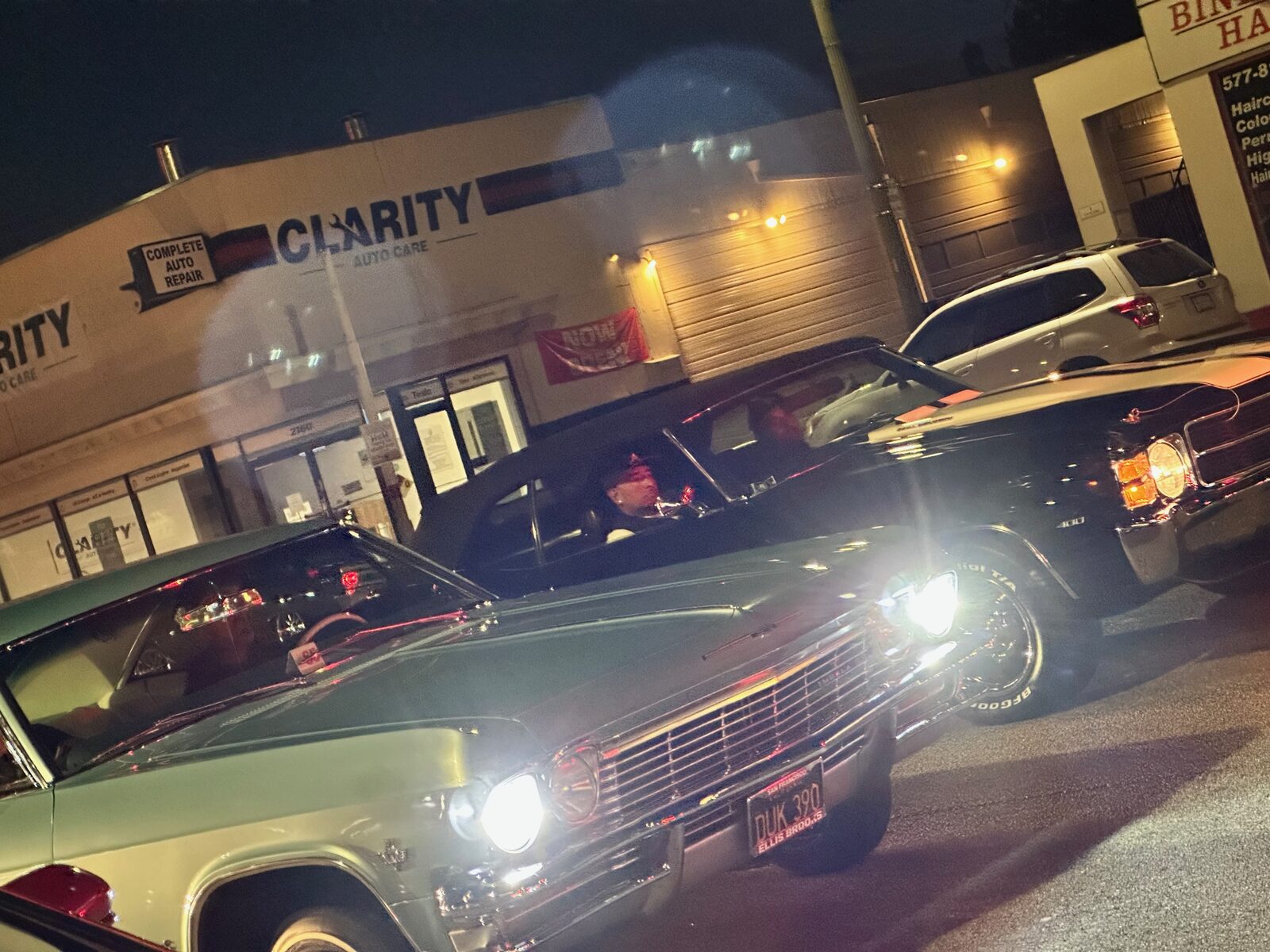 Classic cars with headlights on at night outside a lit storefront