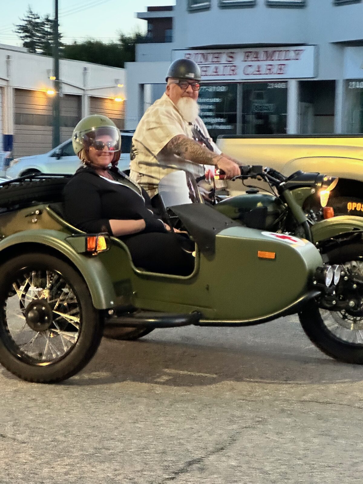 A bearded rider and a passenger in the sidecar of an olive green motorcycle
